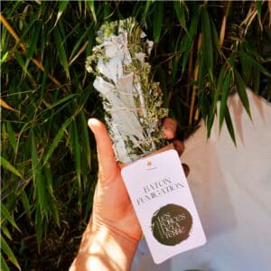 Hand holding a wrapped bundle of dried herbs tied with twine, with a white card and circular logo, against green grass background